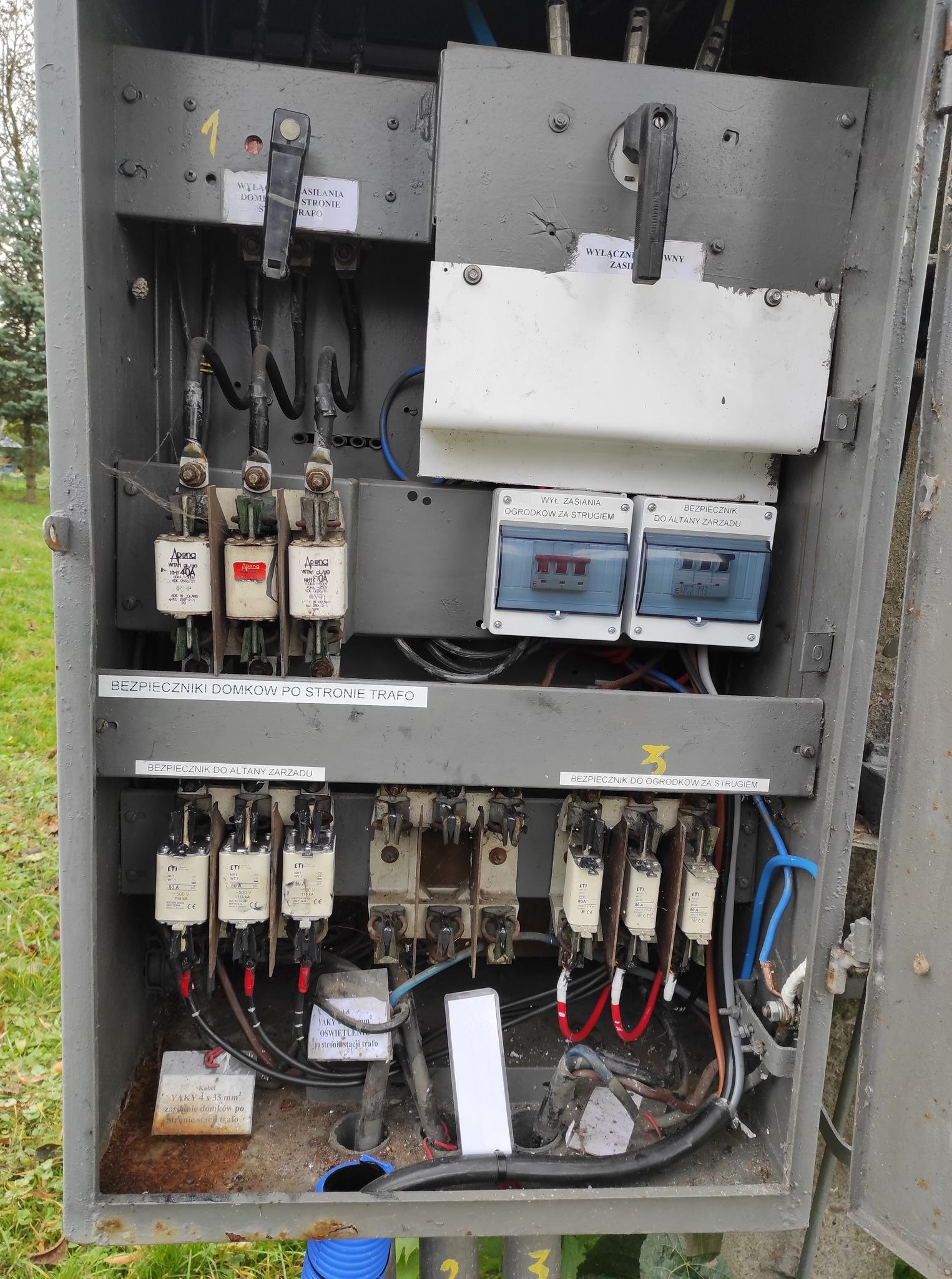 Otwarta skrzynka elektryczna z bezpiecznikami, wyłącznikami i kablami, widoczne oznaczenia obwodów i parametry techniczne, szara obudowa z widocznymi śladami użytkowania na zewnątrz.