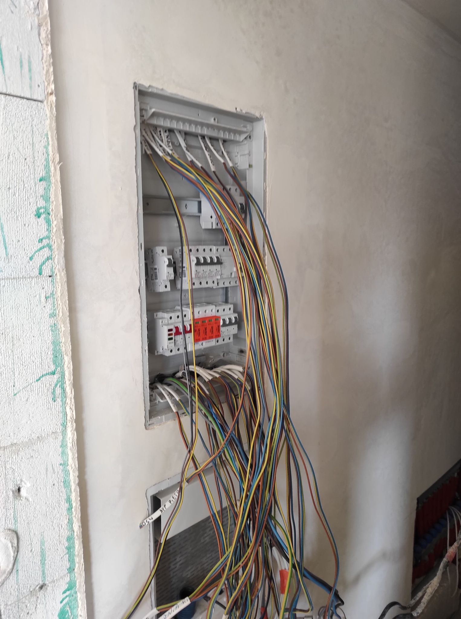 Rozdzielnica elektryczna w trakcie montażu w surowym stanie, widoczne liczne, kolorowe przewody wychodzące z puszki, ściana w trakcie wykańczania.