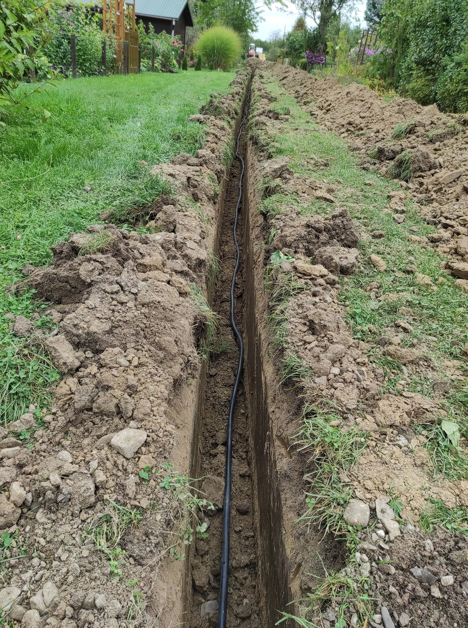 Wąski wykop w trawniku z ułożonym kablem, widok z góry na tle ogrodu z domkiem.