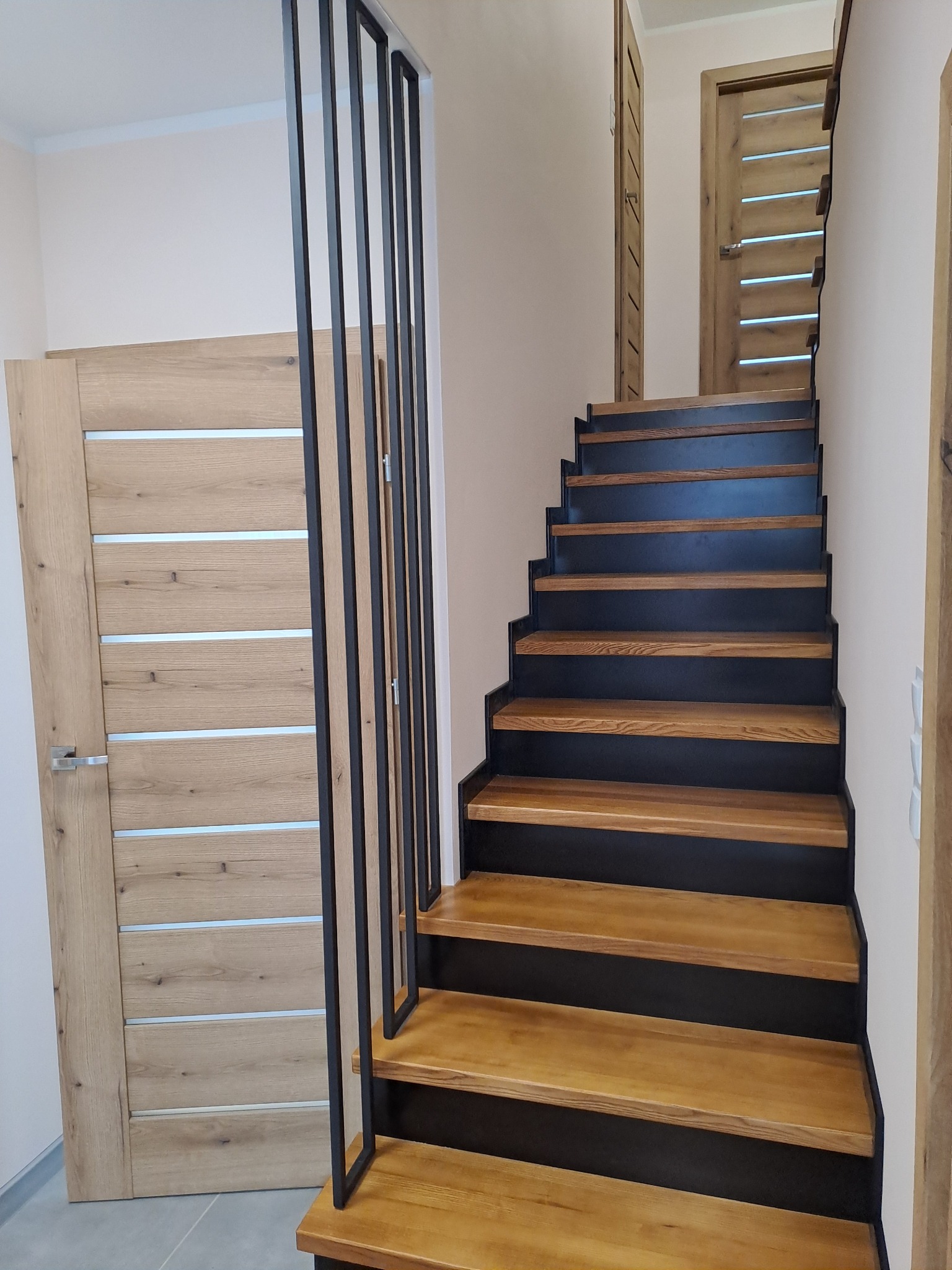 Nowoczesne schody z drewnianymi stopniami i czarną metalową konstrukcją, widoczne drzwi z drewnopodobną okleiną i srebrnymi wstawkami w tle.