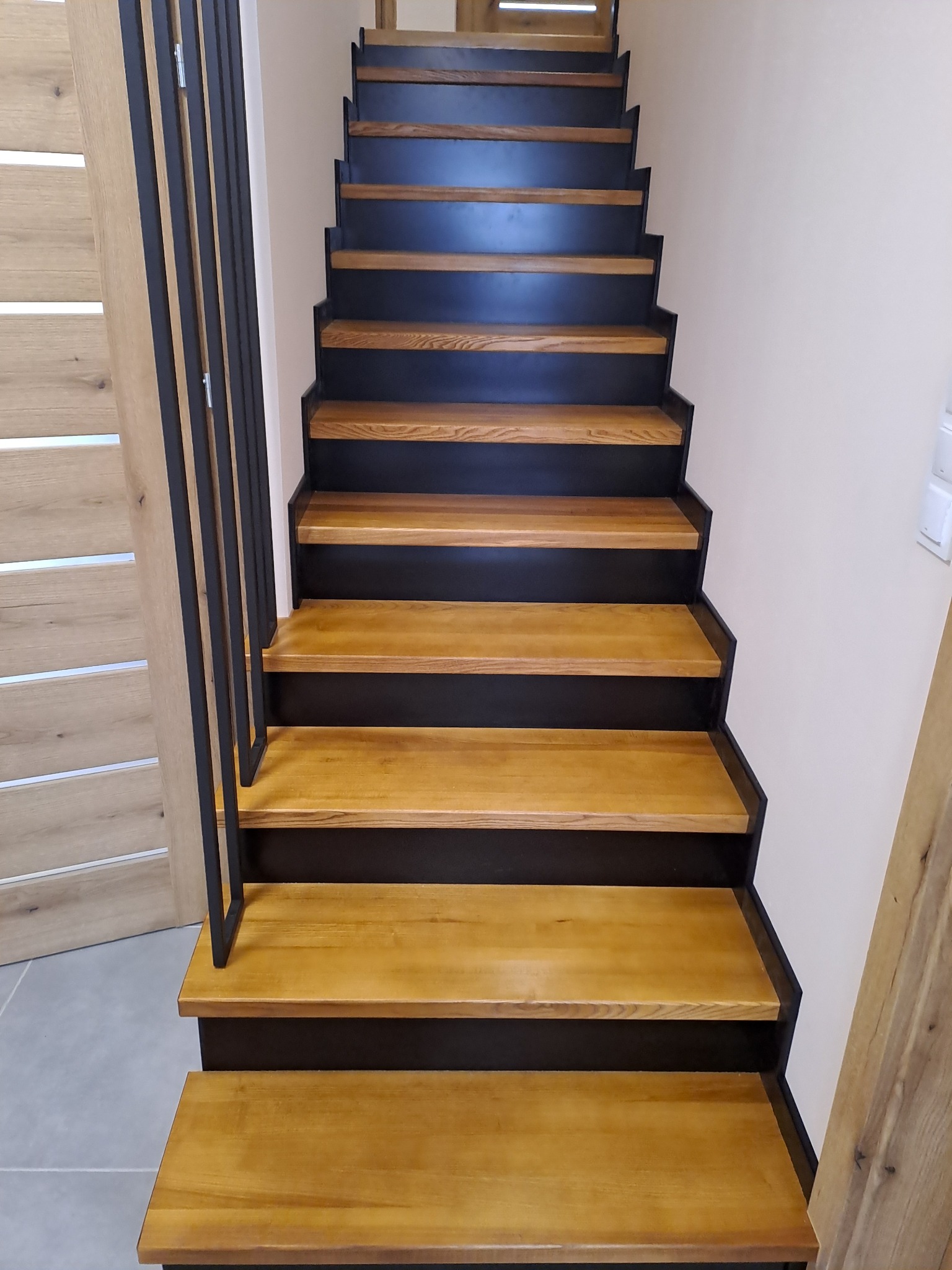 Nowoczesne schody z drewnianymi stopniami i czarną metalową konstrukcją, widok z dołu, obok widoczna drewniana ościeżnica drzwi.