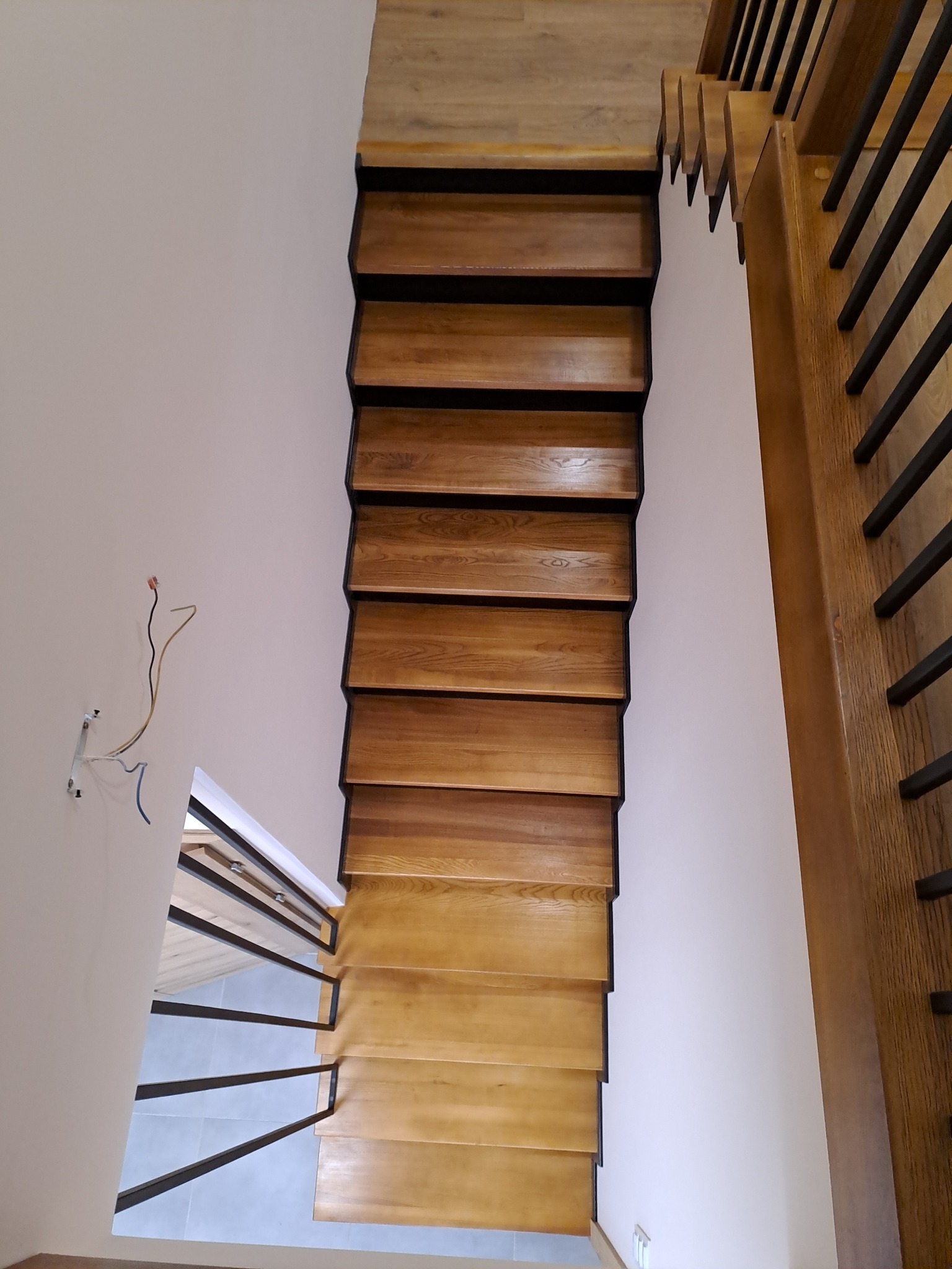 Widok z góry na drewniane schody z czarnymi elementami konstrukcyjnymi, widoczne fragmenty balustrady z metalowymi prętami, na ścianie wystające kable elektryczne.