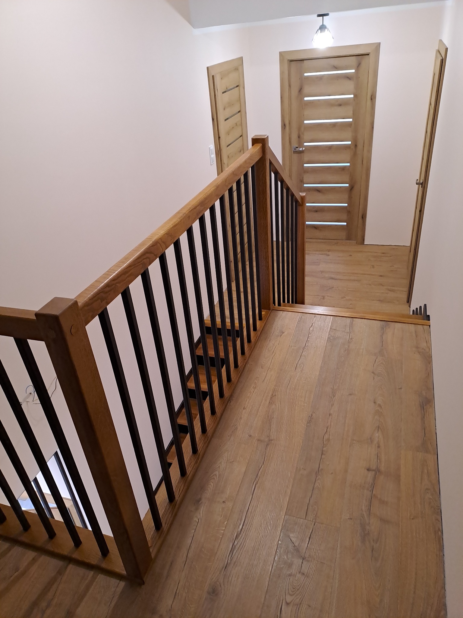 Widok z góry na drewniane schody z czarnymi metalowymi balustradami, prowadzące w dół, z widocznymi drzwiami w tle i drewnianą podłogą imitującą panele.