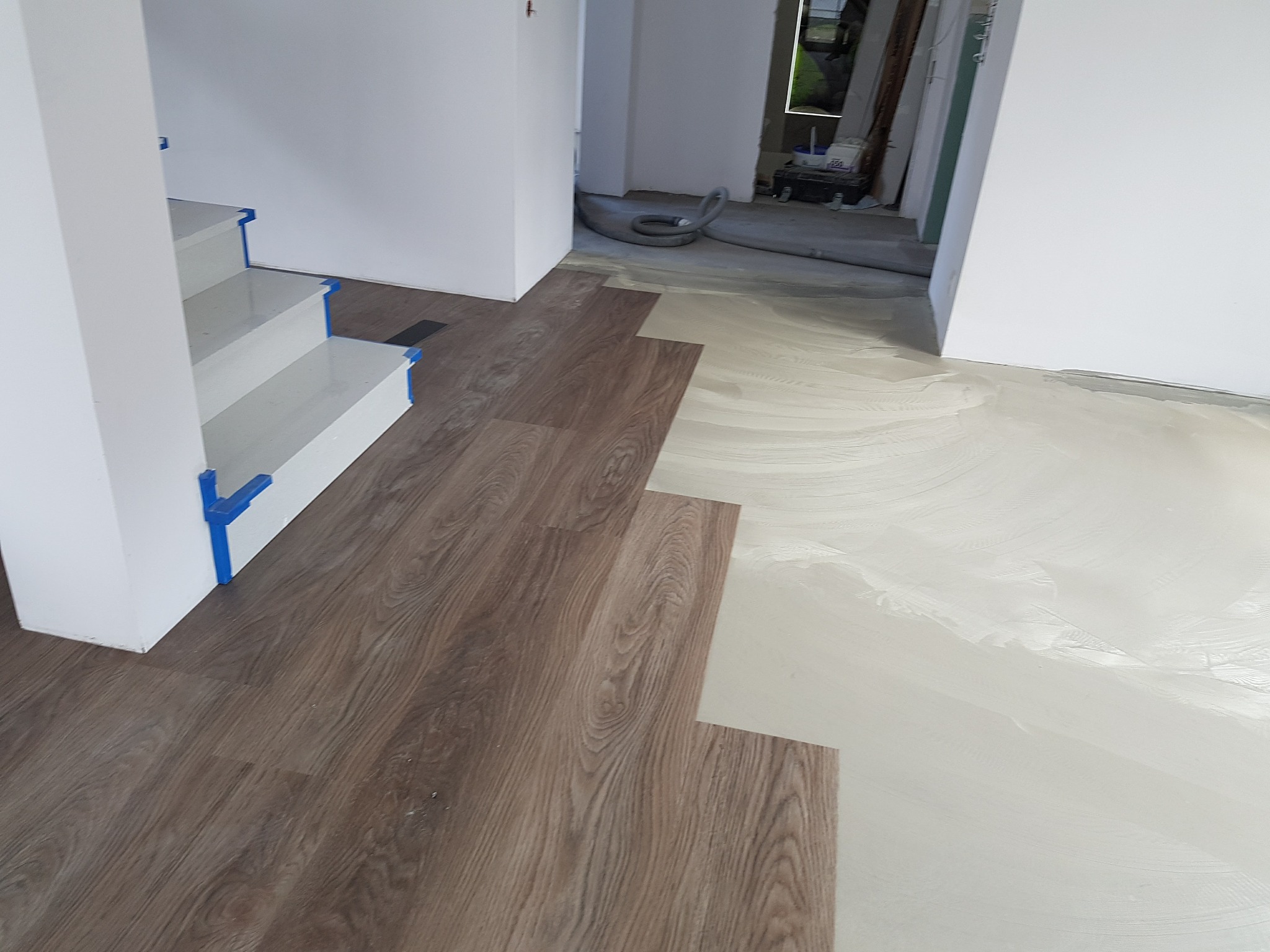 Układanie paneli podłogowych w jasnym wnętrzu, widoczny fragment schodów zabezpieczony niebieską taśmą malarską, podłoże przygotowane pod montaż.