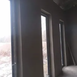 Świeżo otynkowane ściany w surowym wnętrzu z trzema oknami, widoczny surowy betonowy strop i fragment podłogi, na zewnątrz widok na zimowy krajobraz.