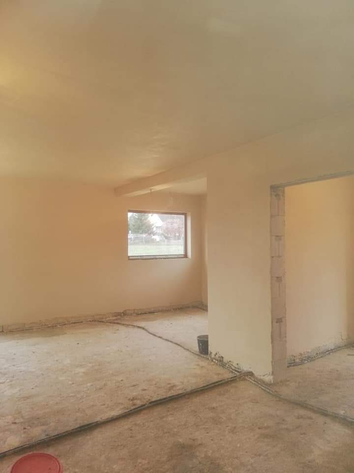 Pokój po nałożeniu tynków maszynowych, widoczne przewody na podłodze, okno z widokiem na zewnątrz i niedokończone obramowanie drzwi.