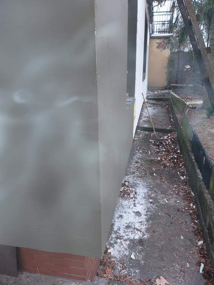 Świeżo otynkowana ściana budynku z widocznymi śladami po zacieraniu, zewnętrzna elewacja w trakcie prac wykończeniowych, widoczny fragment ceglanej podstawy, liście na ziemi.