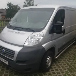 Srebrny Fiat Ducato, bus transportowy, zaparkowany na kostce brukowej przed posesją z niskim ogrodzeniem.