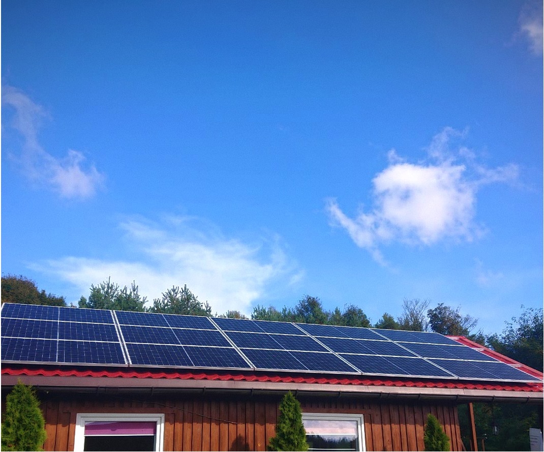 Panele fotowoltaiczne na brązowym dachu domu jednorodzinnego, widok z dołu, błękitne niebo z chmurami w tle.