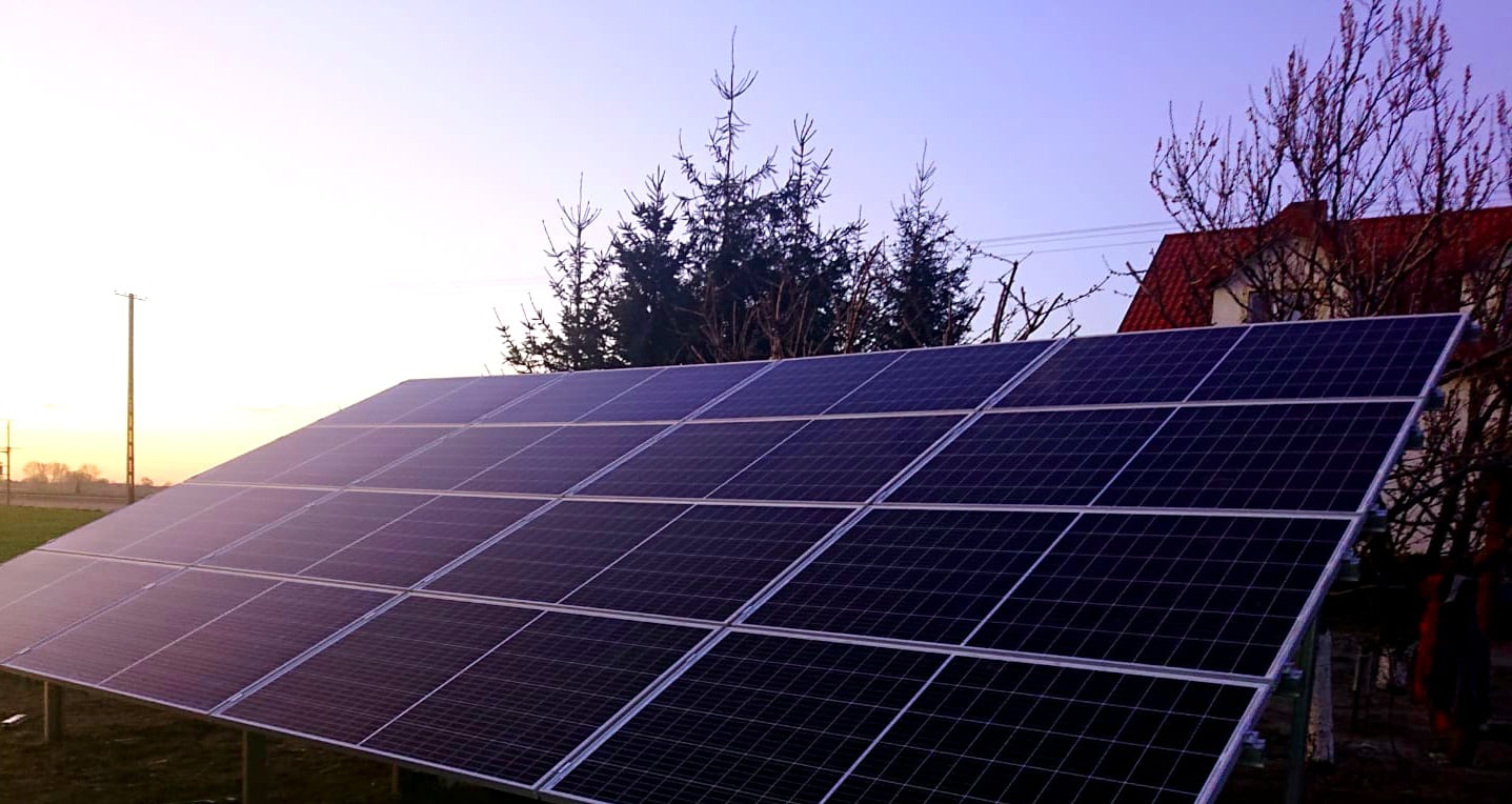 Panel słoneczny na metalowej konstrukcji, z widocznym domem z czerwoną dachówką w tle, oświetlony wschodzącym słońcem.