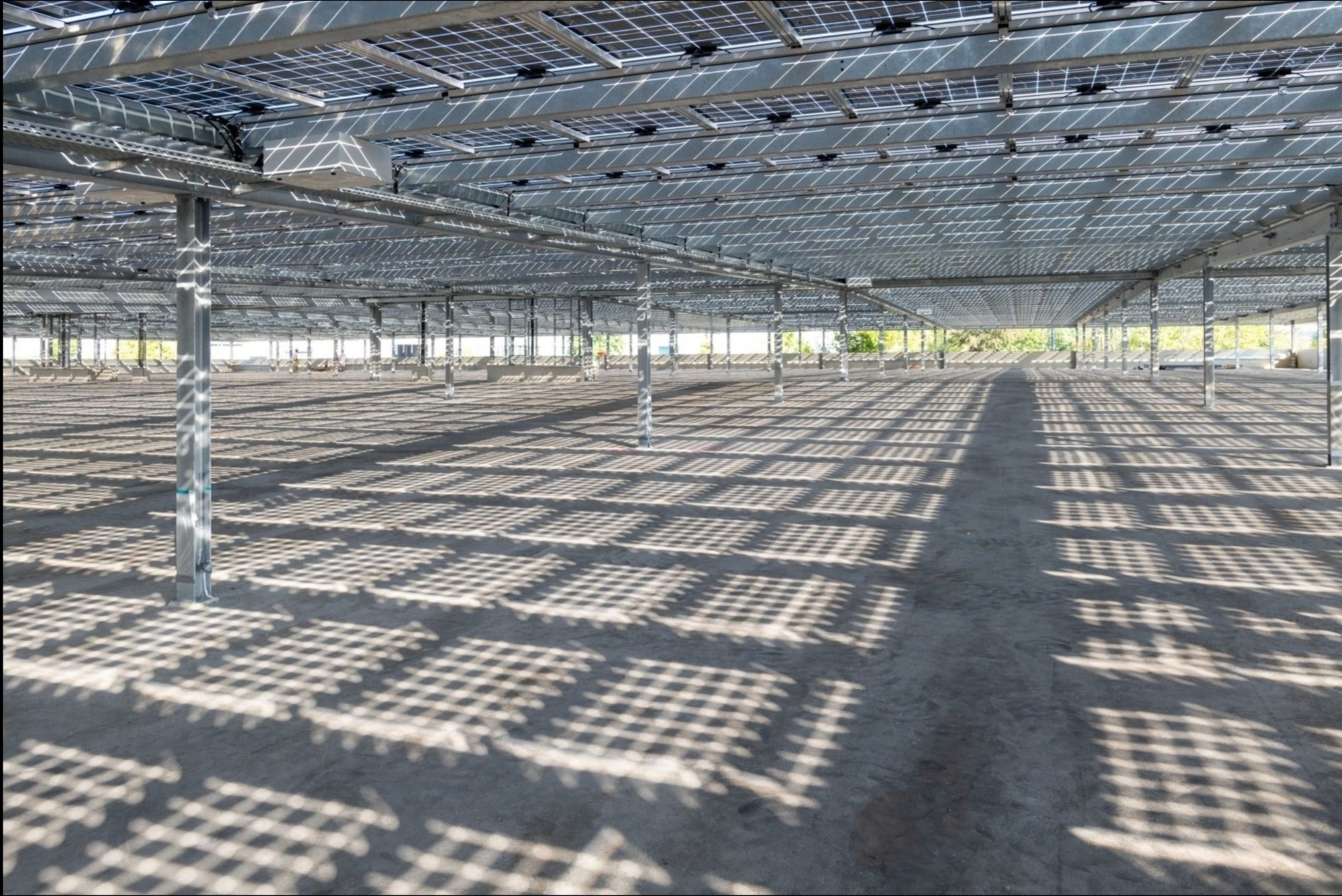 Rozległa instalacja paneli słonecznych na konstrukcji wsporczej, rzucająca geometryczne cienie na betonową posadzkę hali.