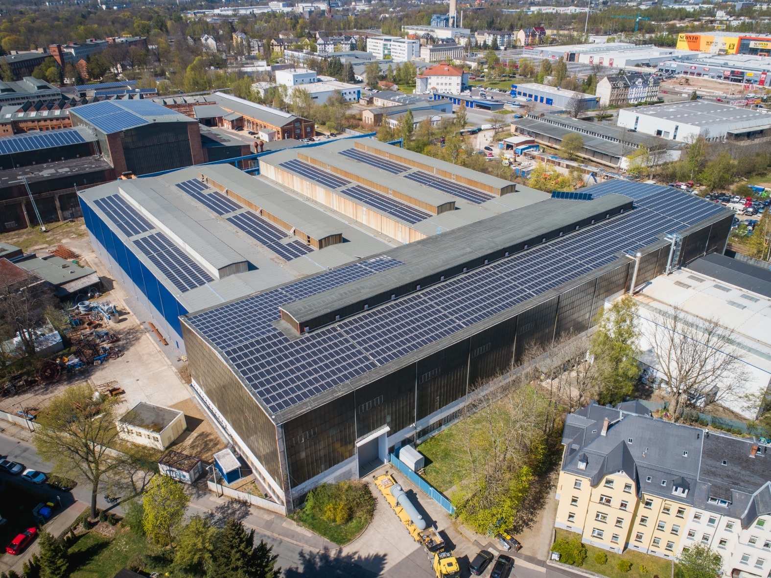 Widok z lotu ptaka na duży budynek przemysłowy z zainstalowanymi panelami słonecznymi na dachu, otoczony zielenią i innymi budynkami.