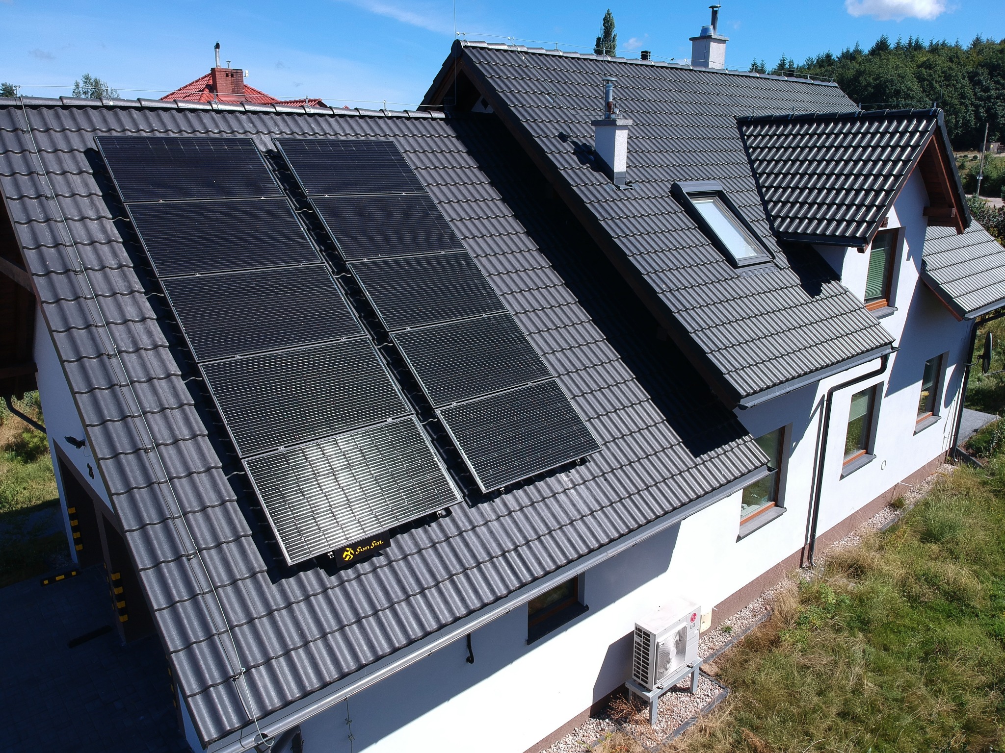 Cztery czarne panele słoneczne na ciemnoszarym dachu domu jednorodzinnego z oknem dachowym, widoczne logo producenta na jednym z paneli, obok drzewa i błękitne niebo.