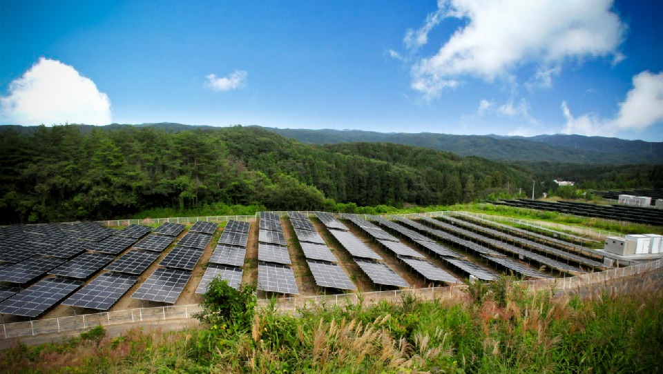 Rozległa farma paneli słonecznych z widokiem na zalesione wzgórza pod błękitnym niebem z chmurami, widoczne ogrodzenie i trawy na pierwszym planie.