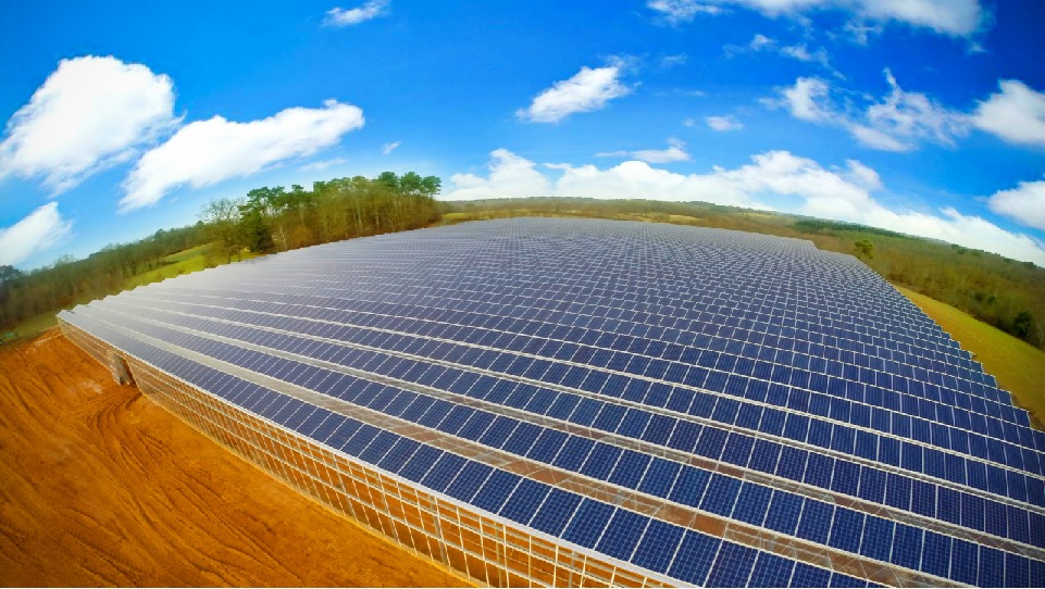 Rozległa farma paneli słonecznych na tle błękitnego nieba z chmurami i zieleni drzew, uchwycona z perspektywy lotniczej.