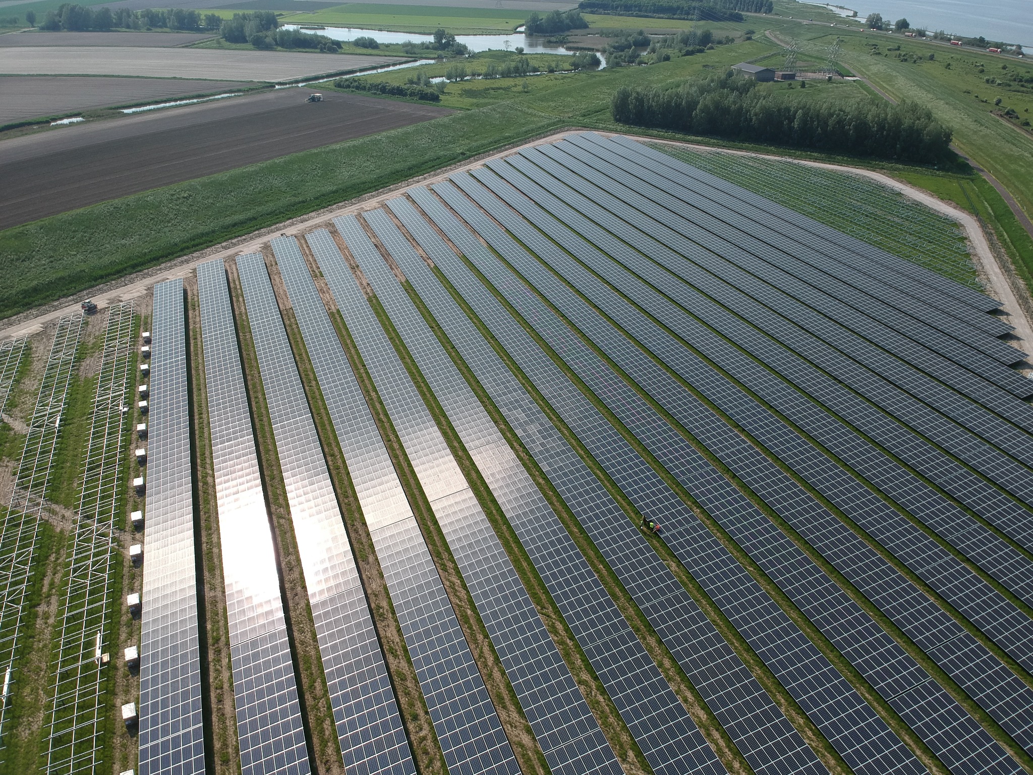 Rozległa farma paneli słonecznych widziana z lotu ptaka, z polami uprawnymi i zbiornikiem wodnym w tle.