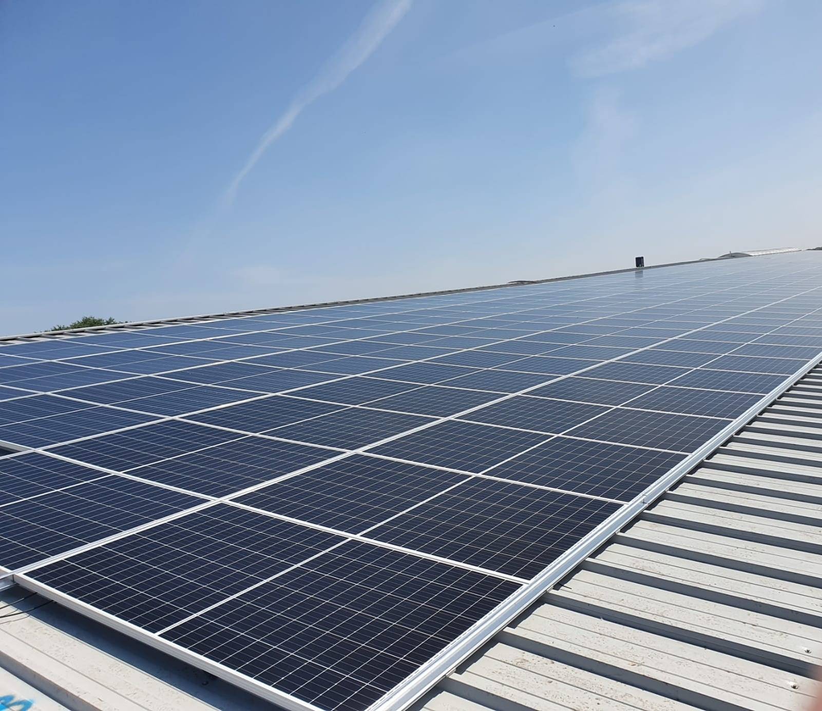 Rząd paneli słonecznych na metalowym dachu w słoneczny dzień, widoczny ślad kondensacji na niebie.