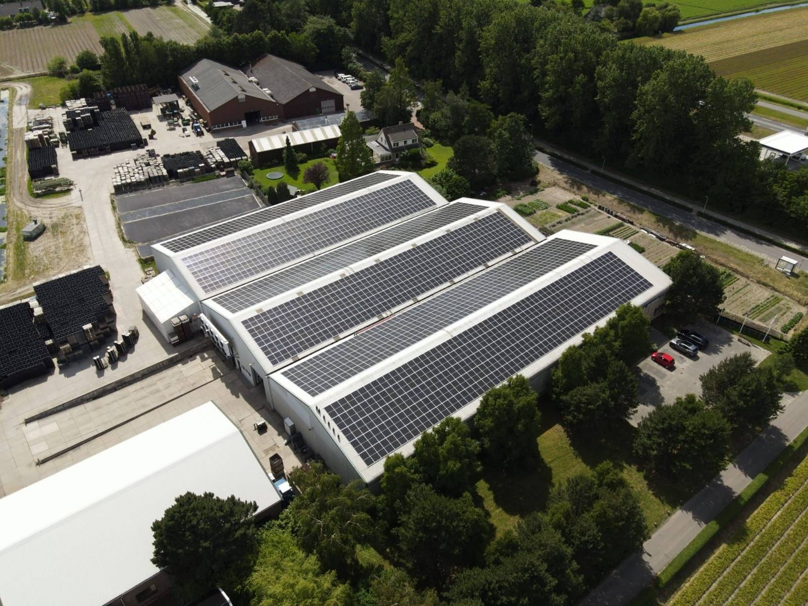 Widok z lotu ptaka na duży budynek z trzema dachami pokrytymi panelami słonecznymi, otoczony zielenią, z zaparkowanymi samochodami w pobliżu.