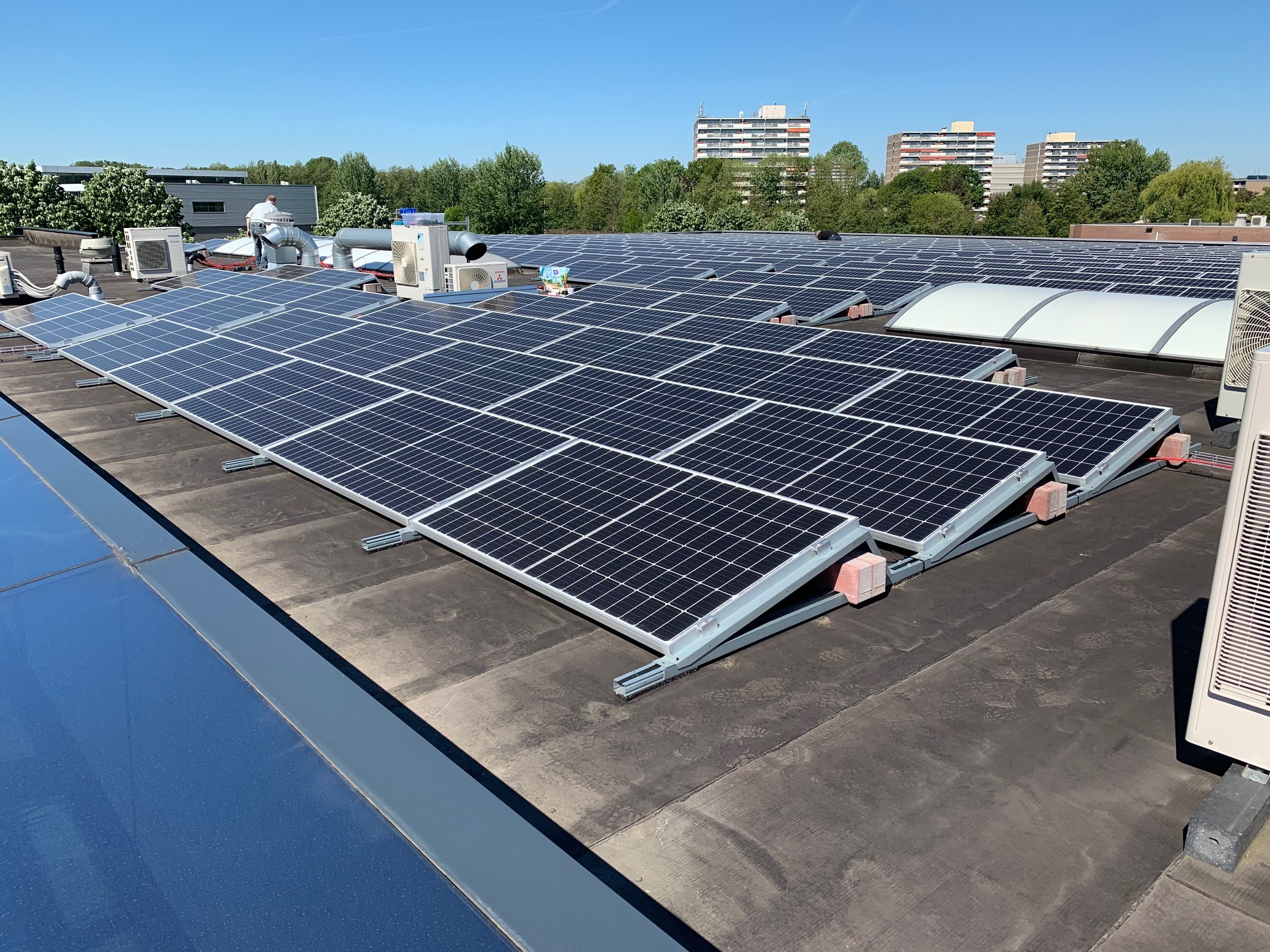 Rozległa instalacja fotowoltaiczna na płaskim dachu budynku, widoczne panele słoneczne zamontowane na metalowych konstrukcjach, w tle budynki mieszkalne i drzewa, błękitne niebo.
