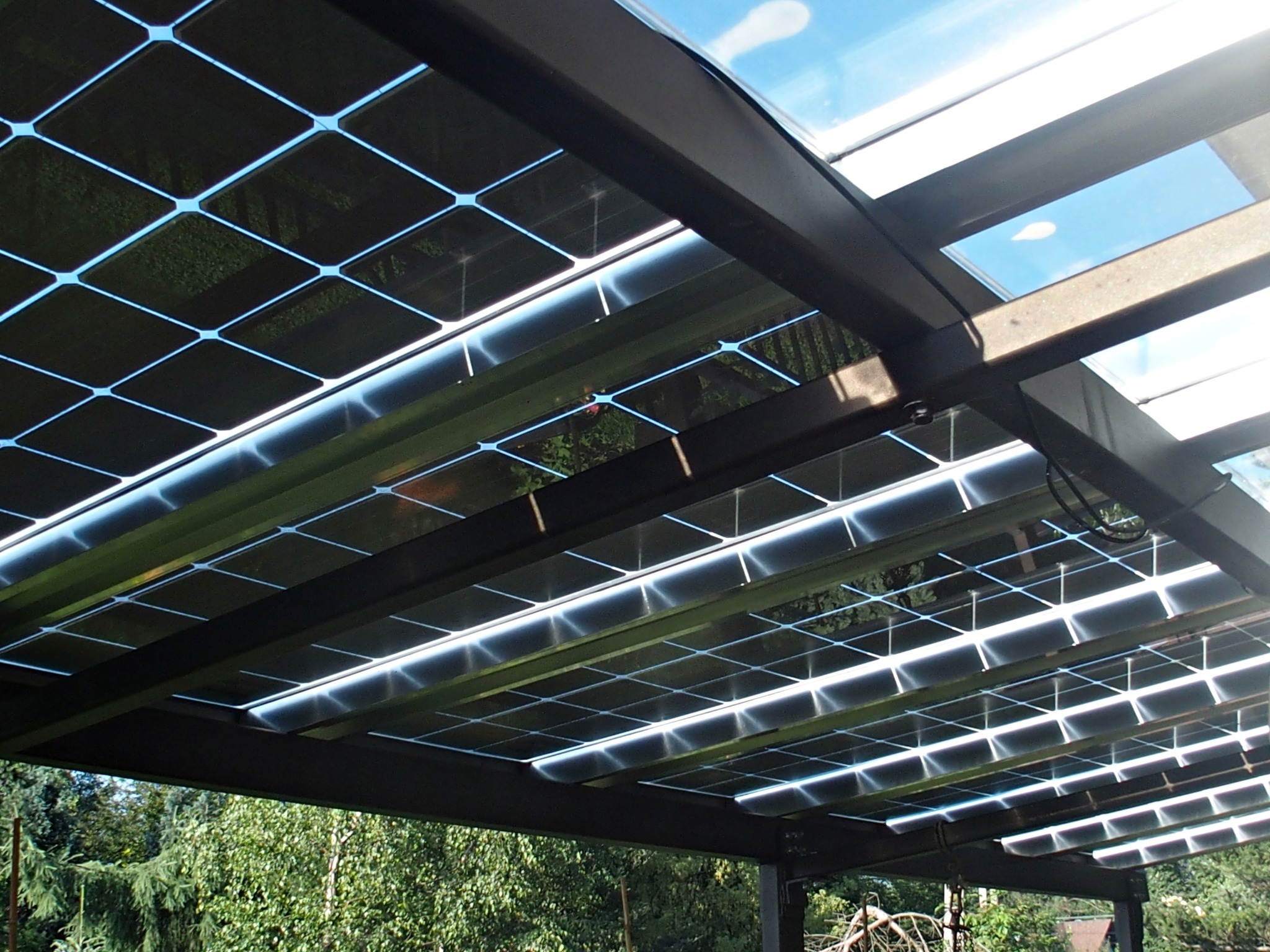 Zadaszenie tarasu zintegrowane z panelami słonecznymi, widok od dołu na tle zieleni i błękitnego nieba.