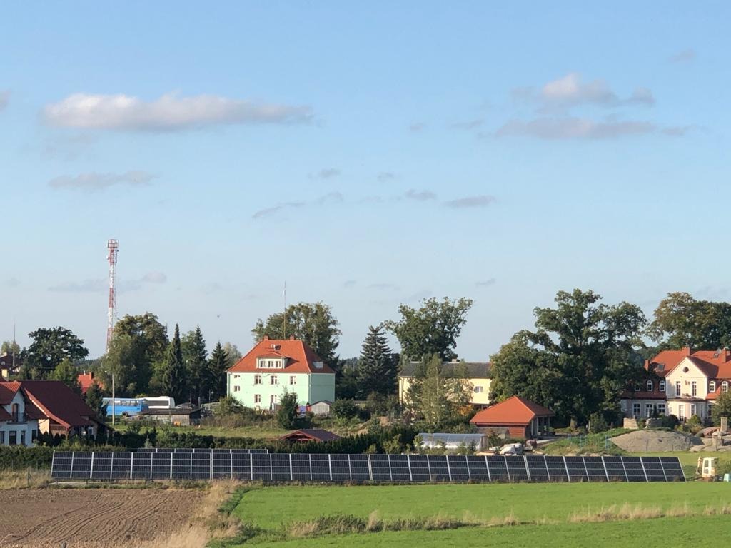 Instalacja paneli słonecznych na polu uprawnym przed zabudowaniami wiejskimi w słoneczny dzień.