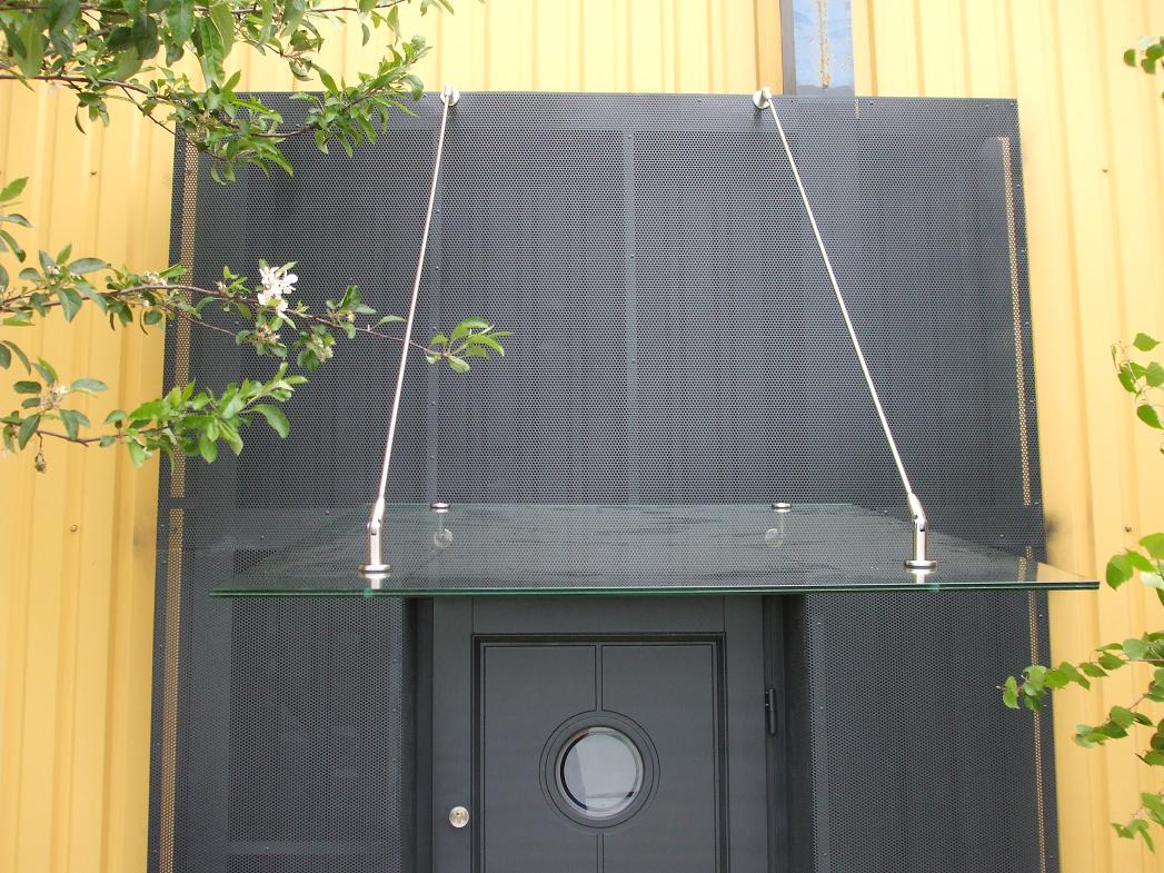 Szklany daszek nad szarymi drzwiami wejściowymi, podtrzymywany przez stalowe linki przymocowane do ażurowej ściany, na tle żółtej blachy falistej i zielonych gałęzi drzew.