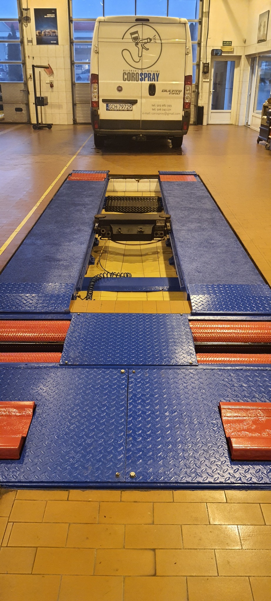 Wnętrze warsztatu samochodowego z podnośnikiem najazdowym, w tle zaparkowany biały bus firmy Corospray z widocznym logo i danymi kontaktowymi.