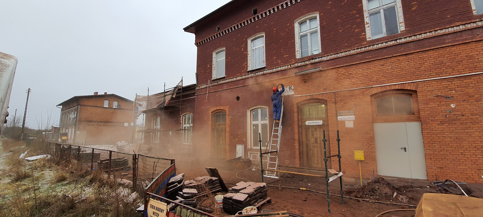 Pracownik w kombinezonie i kasku na drabinie, oczyszcza ceglaną elewację starego dworca kolejowego za pomocą strumienia ściernego, widoczne rusztowania i sterta kostki brukowej.