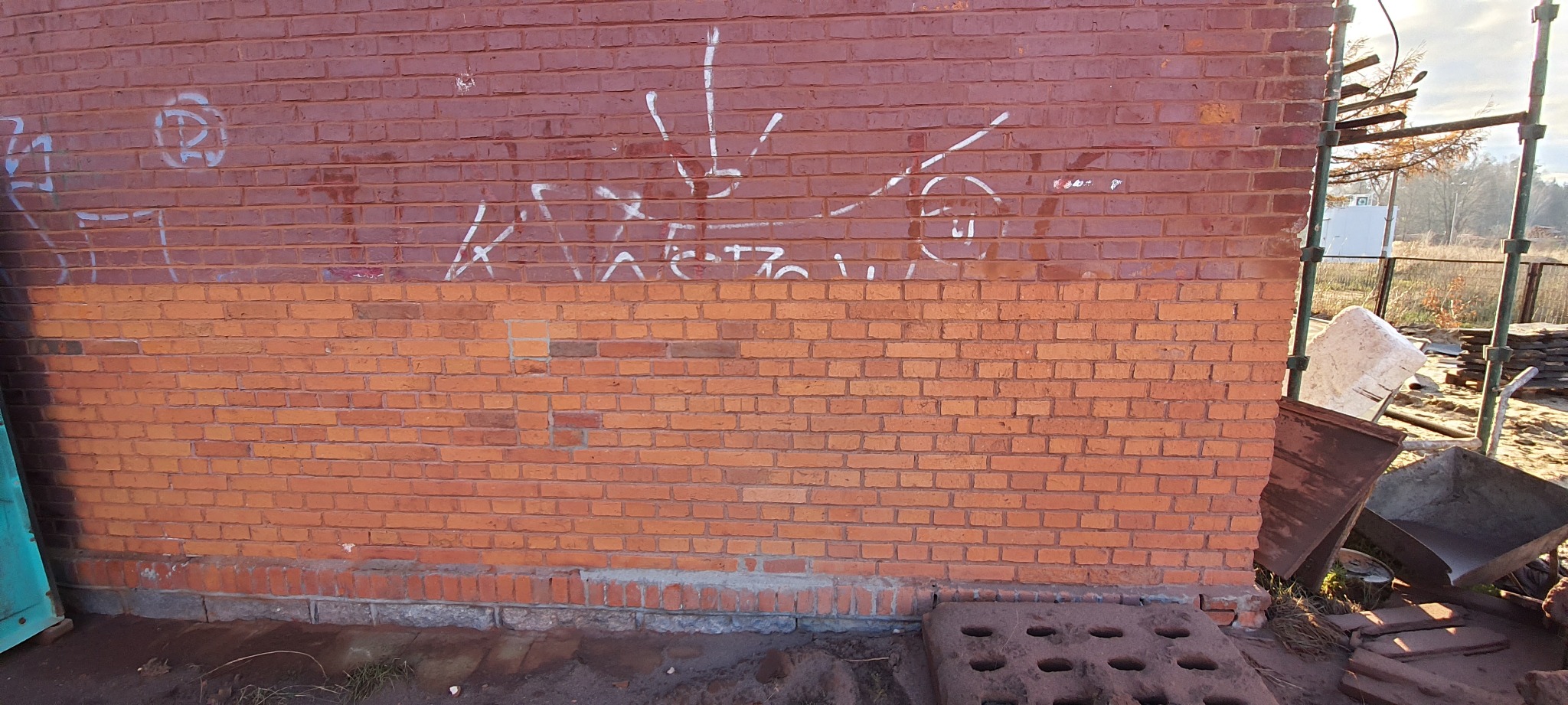 Ceglana ściana budynku z częściowo usuniętą farbą i graffiti, widoczne ślady po piaskowaniu, obok rusztowanie i gruz.