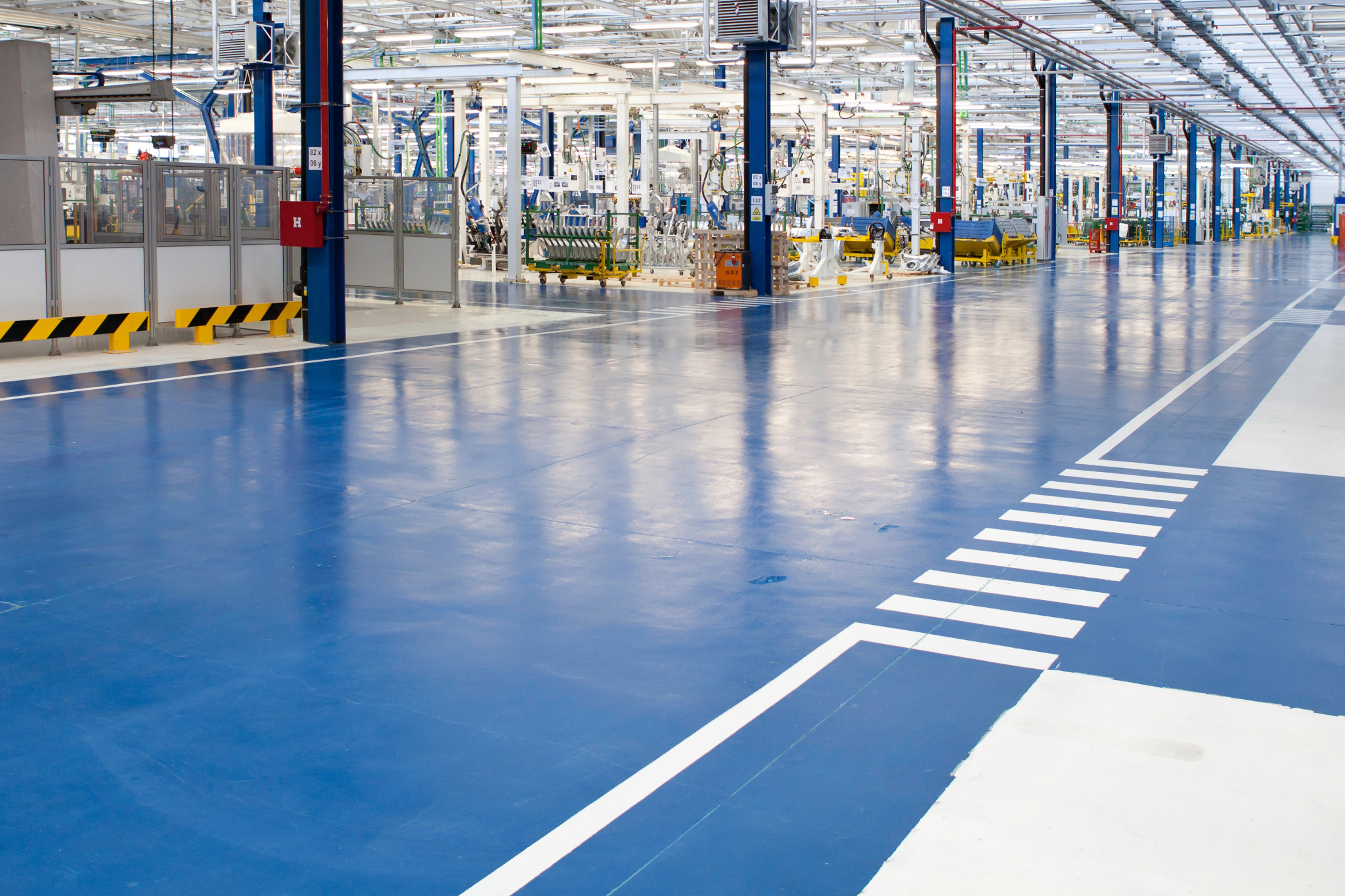 Wnętrze hali produkcyjnej z czystą, niebieską podłogą i białymi pasami, oświetlonej licznymi lampami sufitowymi, z widocznymi elementami linii produkcyjnej i instalacjami technicznymi.