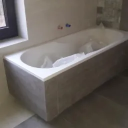 Nowoczesna wanna obudowana płytkami imitującymi drewno, częściowo zabezpieczona folią, zamontowana w łazience w Górze Kalwarii, z widocznymi przyłączami wodnymi.