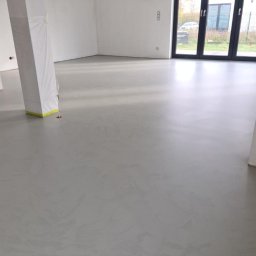 Podłogi żywiczne, mikrocement Epoxyfloor - Szara, gładka posadzka w surowym wnętrzu z kolumną zabezpieczoną folią malarską, widok na okno tarasowe.