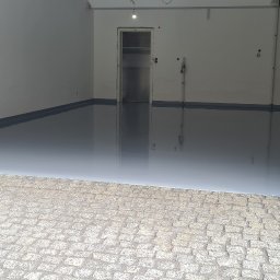 Podłogi żywiczne, mikrocement Epoxyfloor - Błyszcząca, szara podłoga w garażu z wejściem wyłożonym kostką brukową i ceglanymi filarami po bokach wjazdu. W tle widoczne drzwi do pomieszczenia.