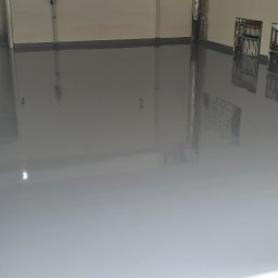 Podłogi żywiczne, mikrocement Epoxyfloor - Gładka, szara podłoga w garażu, świeżo po wylaniu, z widocznymi instalacjami elektrycznymi na ścianie i fragmentem ceglanej ściany po lewej stronie.