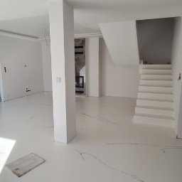 Podłogi żywiczne, mikrocement Epoxyfloor - Biała posadzka z subtelnym wzorem żyłkowania w nowoczesnym wnętrzu domu z białymi ścianami, schodami i dużym oknem na ogród.
