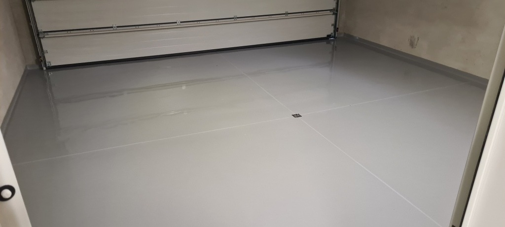 Szara, gładka podłoga w garażu z widocznym odpływem podłogowym i częściowo otwartą bramą garażową.