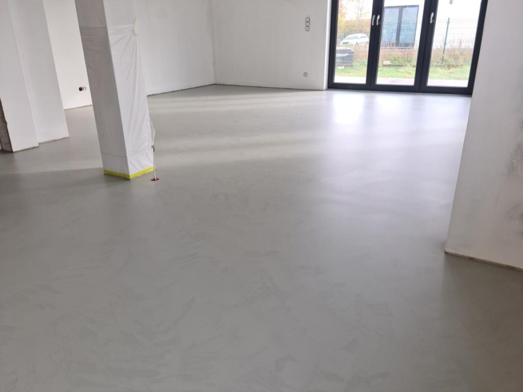 Szara, gładka posadzka w surowym wnętrzu z kolumną zabezpieczoną folią malarską, widok na okno tarasowe.