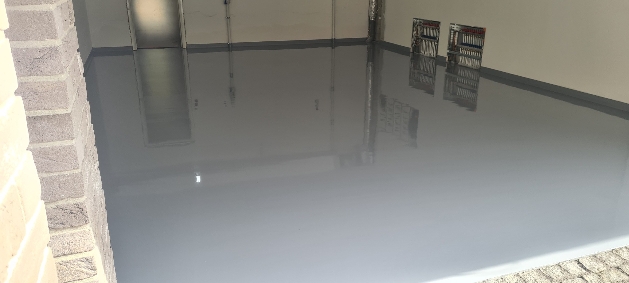 Gładka, szara podłoga w garażu, świeżo po wylaniu, z widocznymi instalacjami elektrycznymi na ścianie i fragmentem ceglanej ściany po lewej stronie.