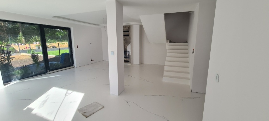 Biała posadzka z subtelnym wzorem żyłkowania w nowoczesnym wnętrzu domu z białymi ścianami, schodami i dużym oknem na ogród.