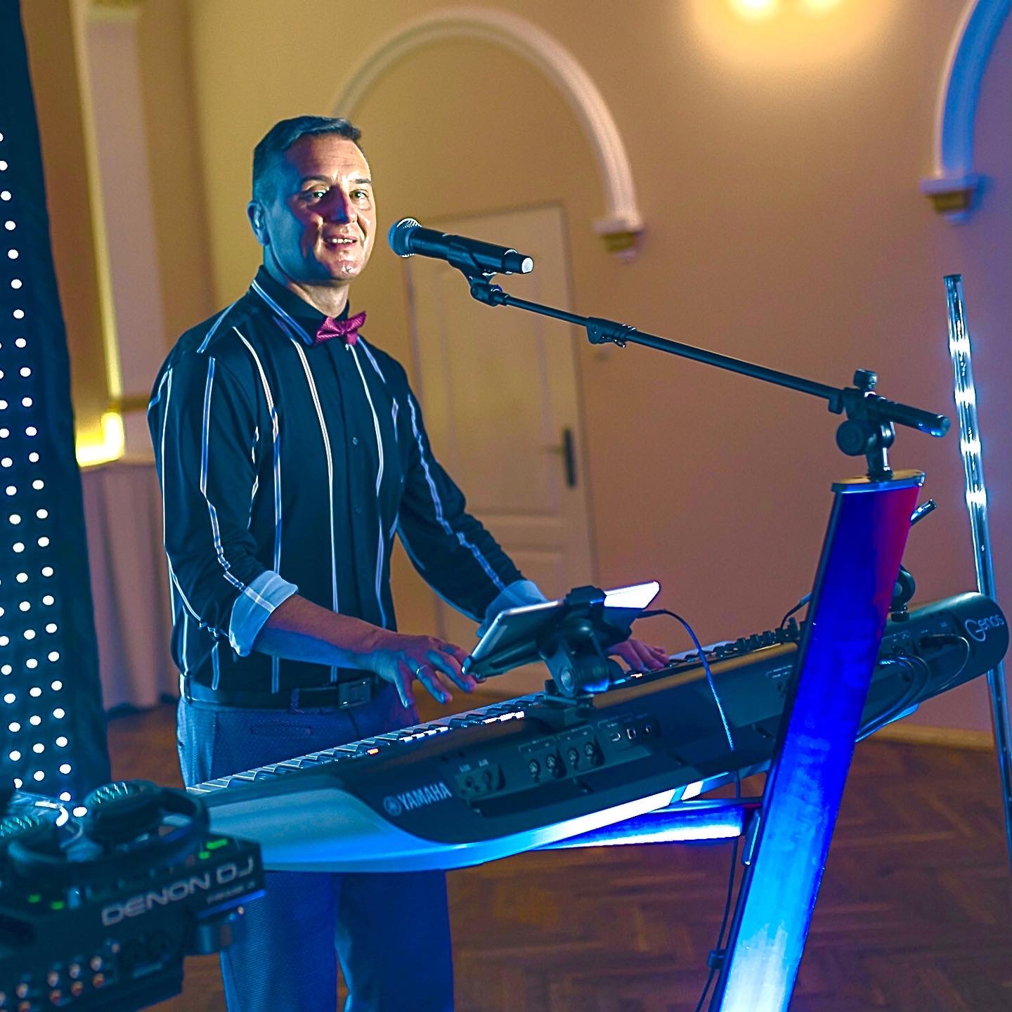 Mężczyzna w koszuli w paski i muszce gra na keyboardzie Yamaha Genos i śpiewa do mikrofonu podczas występu na tle ściany z dekoracyjnymi łukami i oświetleniem LED.