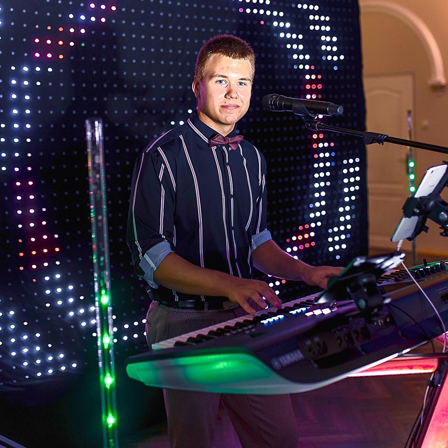 Młody mężczyzna w koszuli w paski i muszce grający na keyboardzie Yamaha na tle ściany z diodami LED, widoczny mikrofon i tablet na statywie.