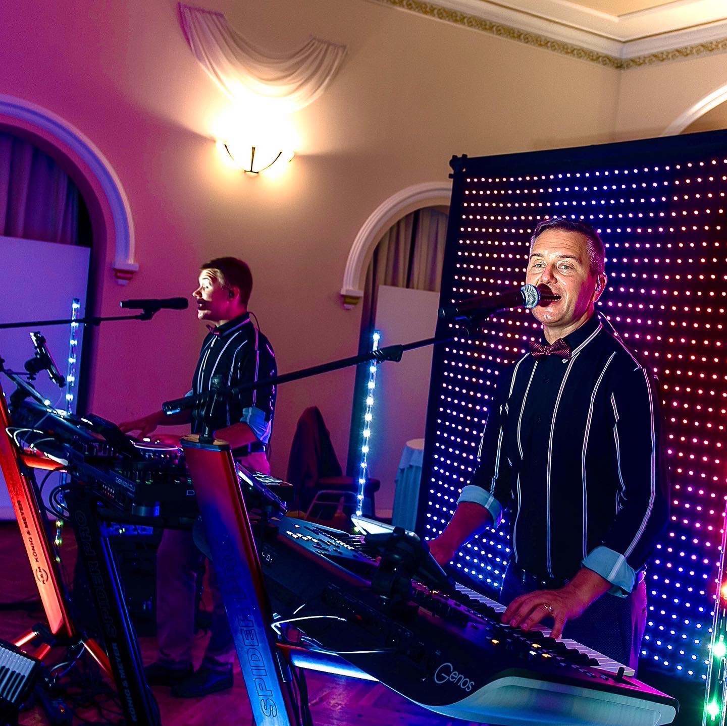 Zespół muzyczny grający na keyboardach Genos podczas występu na tle ściany z diodami LED, oświetlony światłem w odcieniach różu i fioletu. Jeden z muzyków śpiewa do mikrofonu.