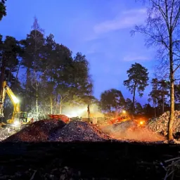 Nocne ujęcie placu budowy w lesie, z żółtą koparką i gruzem podświetlonym światłami, sugerujące prace ziemne i porządkowe po wyburzeniach.