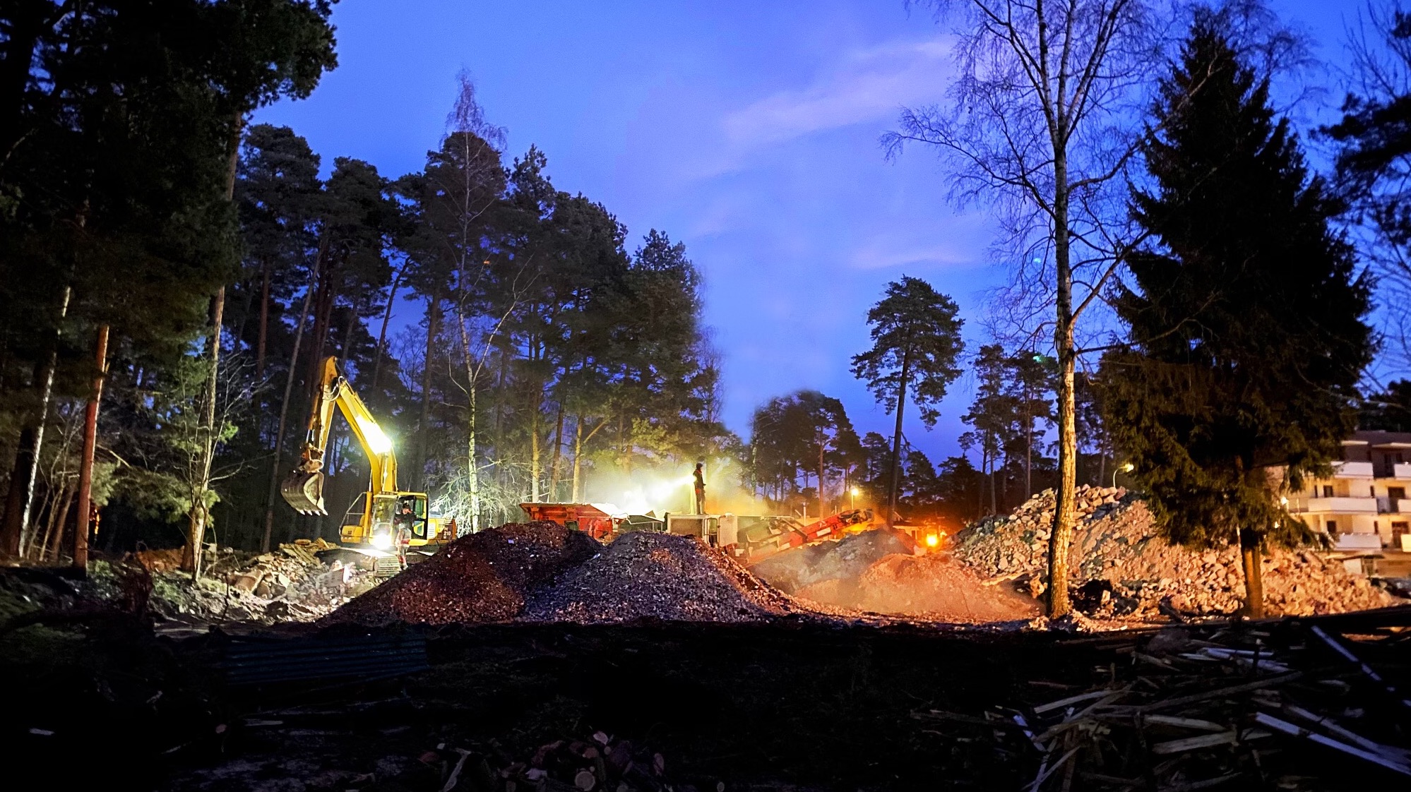 Nocne ujęcie placu budowy w lesie, z żółtą koparką i gruzem podświetlonym światłami, sugerujące prace ziemne i porządkowe po wyburzeniach.