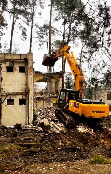 Żółta koparka Hyundai 210LC-9 z uniesioną łyżką podczas rozbiórki częściowo zburzonego, dwukondygnacyjnego budynku z betonu, na tle drzew.