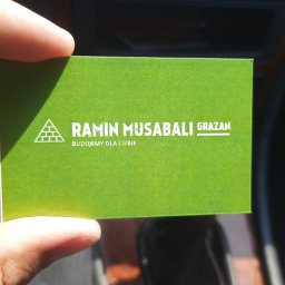 Grazan Spolka Zoo - Zielona wizytówka firmy budowlanej Ramin Musabali Grazan z logo w kształcie piramidy ze stylizowanych cegieł, trzymana w dłoni na tle deski rozdzielczej samochodu.