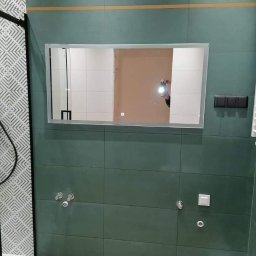 Grazan Spolka Zoo - Wykończona łazienka z zielonymi płytkami, podświetlanym lustrem z wbudowaną szafką i widocznymi przyłączami wodnymi przed montażem armatury.