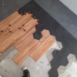 Grazan Spolka Zoo - Układanie podłogi z płytek drewnopodobnych i heksagonalnych, z użyciem pomarańczowych klinów poziomujących; widoczna stopa robotnika w trampku.