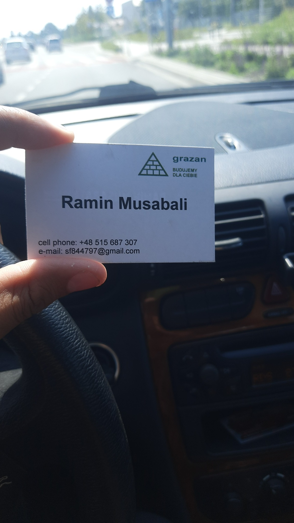 Wizytówka firmy budowlanej Grażan, z logo przedstawiającym mur z cegieł w kształcie trójkąta, trzymana w dłoni na tle deski rozdzielczej samochodu podczas jazdy.