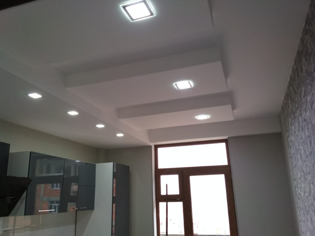 Nowoczesny sufit podwieszany z wbudowanym oświetleniem LED w kuchni, widoczna zabudowa kuchenna w tle, ściana pokryta tapetą o delikatnym wzorze.